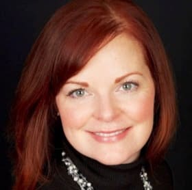 The image is a headshot of a woman with shoulder-length reddish-brown hair. She is smiling and wearing a black turtleneck and a necklace with black and silver details. The background is not visible, focusing the viewer's attention on her. Her appearance is professional, suggesting that this could be a corporate or business-related photograph.