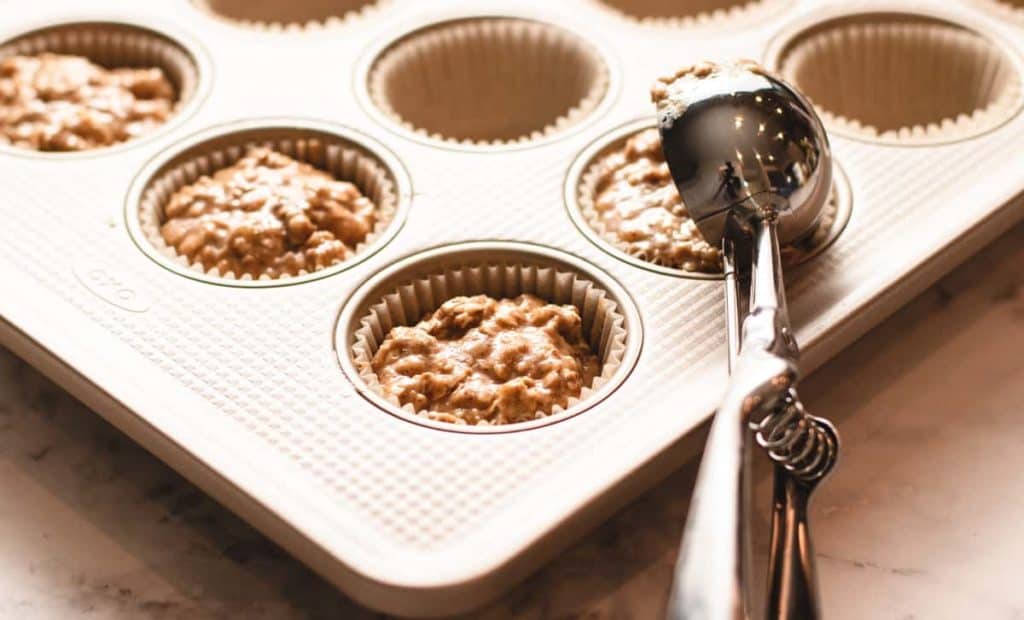 a 12 cup muffin tin with liners filled with batter also shows a ice cream scoop to demonstrate scooping batter into muffin tin