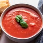 Roasted Tomato Soup in white bowl with basil garnish. A grilled cheese sandwich sits to left of bowl with a spoon on a napkin sits on the right of the bowl.