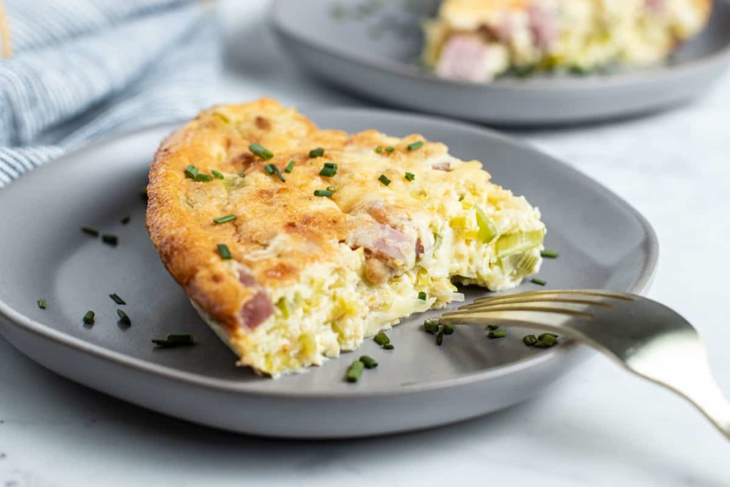 A slice of quiche with ham, cheese, clean and cut leeks, and chives sits on a gray plate with a fork. Another slice is visible in the background, with chopped chives sprinkled on top.
