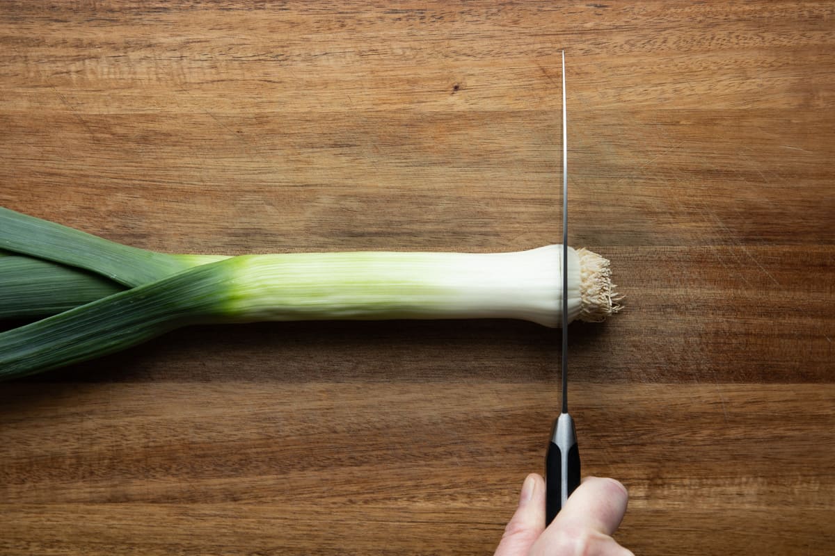 one leek on cutting board with a knife about to cut off the roots leaving the root pad.