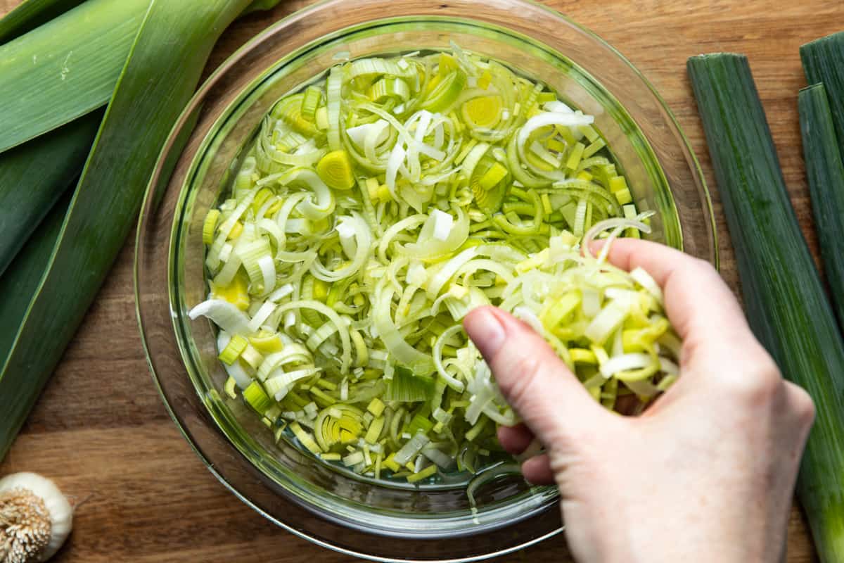 sliced leeks in clear bowl of water with hand agitating them to remove dirt