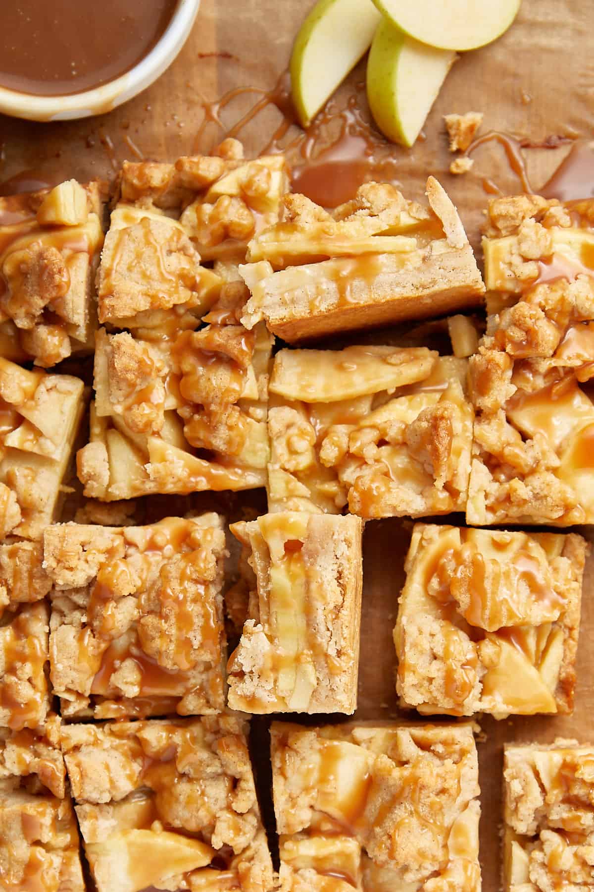 Salted Caramel Apple Pie bars cut into bars on parchment paper with a small bowl of caramel sauce in a white bowl in top left corner. And three apple slices next to bowl. 
