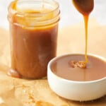 salted caramel sauce dripping into a bowl set on parchment paper. Ball jar sits behind bowl filled with sauce.