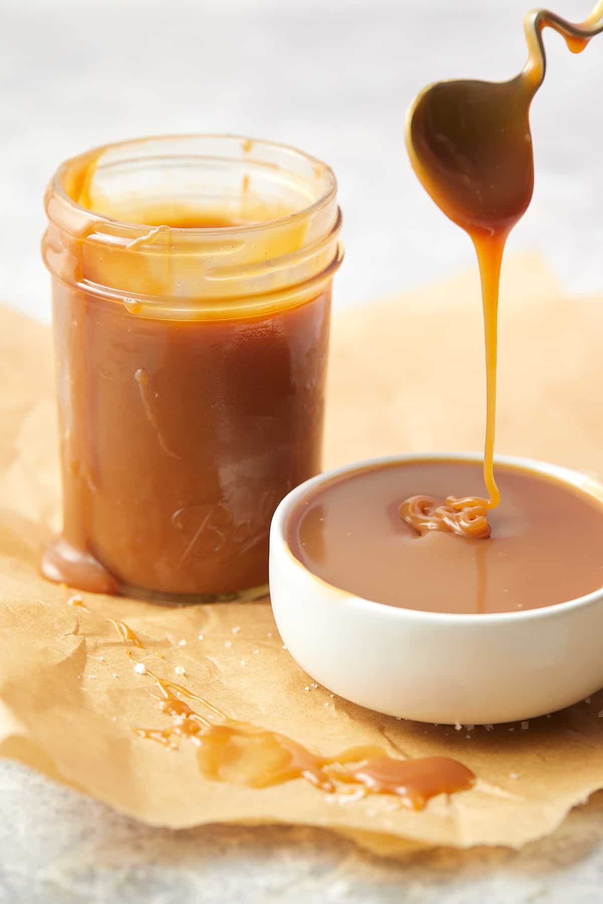 easy homemade salted caramel sauce dripping from a spoon into a small white bowl set on parchment paper. A glass jar filled with caramel sauce sits behind the bowl.