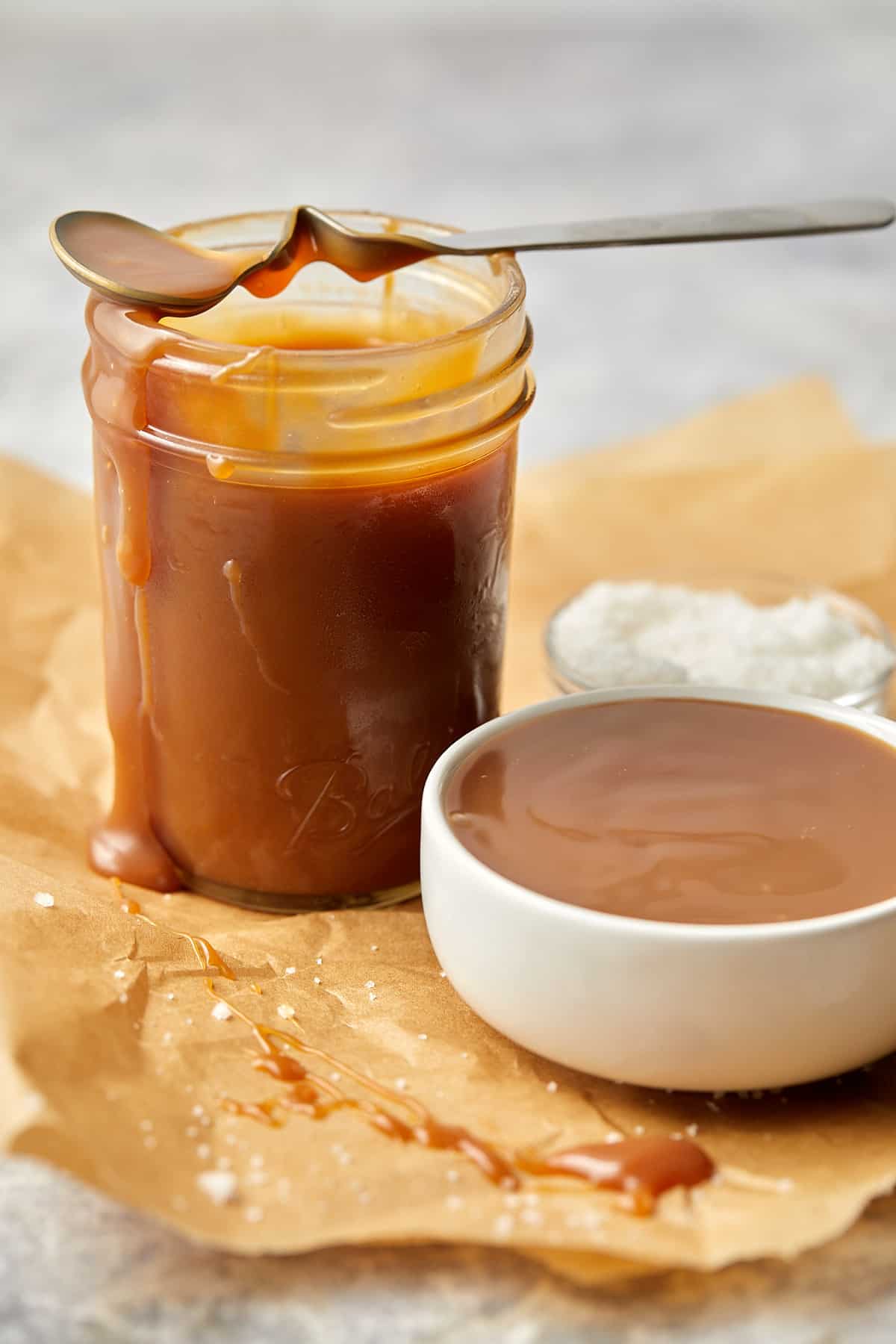 easy homemade salted caramel in small white bowl along with a glass jar filled with sauce. On top of the jar sits a spoon dripping with sauce. Next to the jar sits a small glass bowl of salt. 