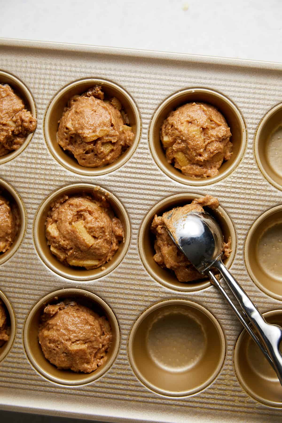 a muffin tin with the batter being poured into cups with a cookie scoop.