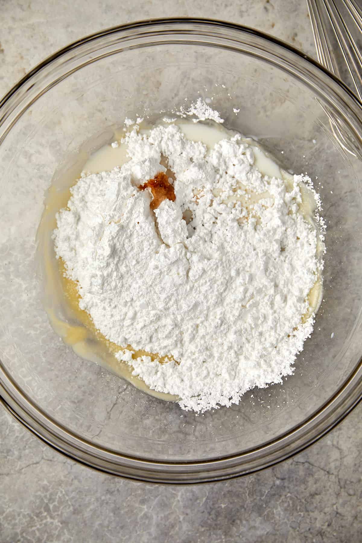 The image shows a clear glass mixing bowl on a gray surface. Inside the bowl, you can see a mound of powdered sugar on top of melted chocolate. Vanilla is also in the mound of powdered sugar. 