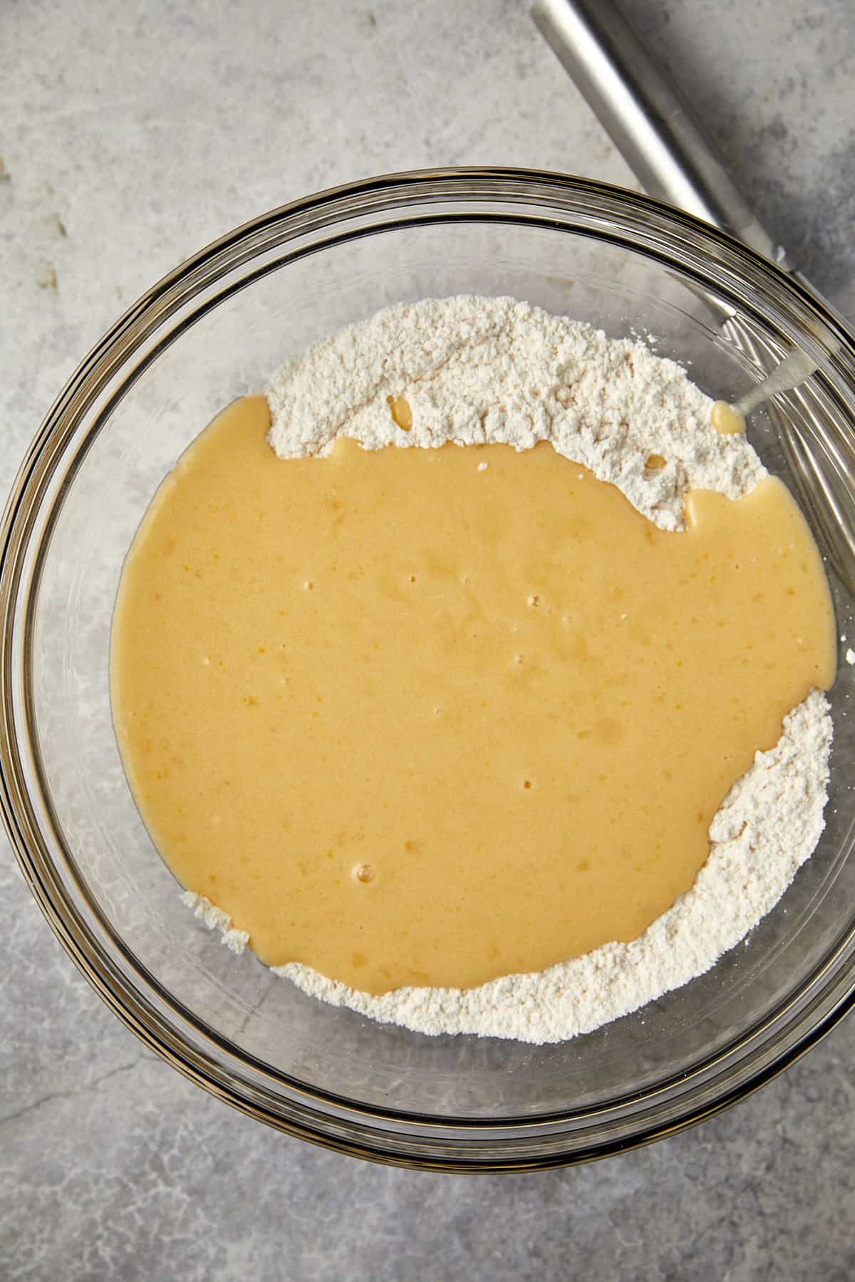 The image shows a glass mixing bowl with a mixture of wet and dry ingredients on a gray surface. One side of the bowl contains a creamy, tan-colored liquid mixture, while the other holds a white flour pile. The ingredients are on the verge of being mixed, indicating a step in a baking recipe where the wet and dry components are combined to form a batter.