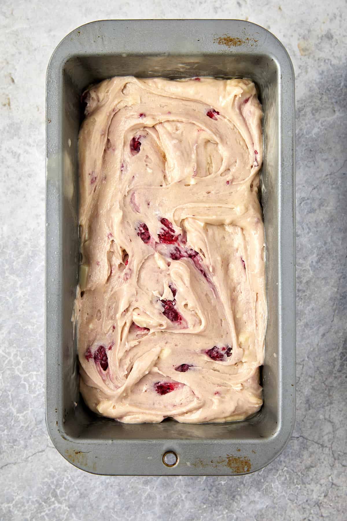 The image shows a rectangular metal baking pan, 9x5 inches, filled with a swirled raspberry and white chocolate batter. The batter is evenly spread within the pan, which sits on a gray surface. The pan appears well-used, with signs of wear. 