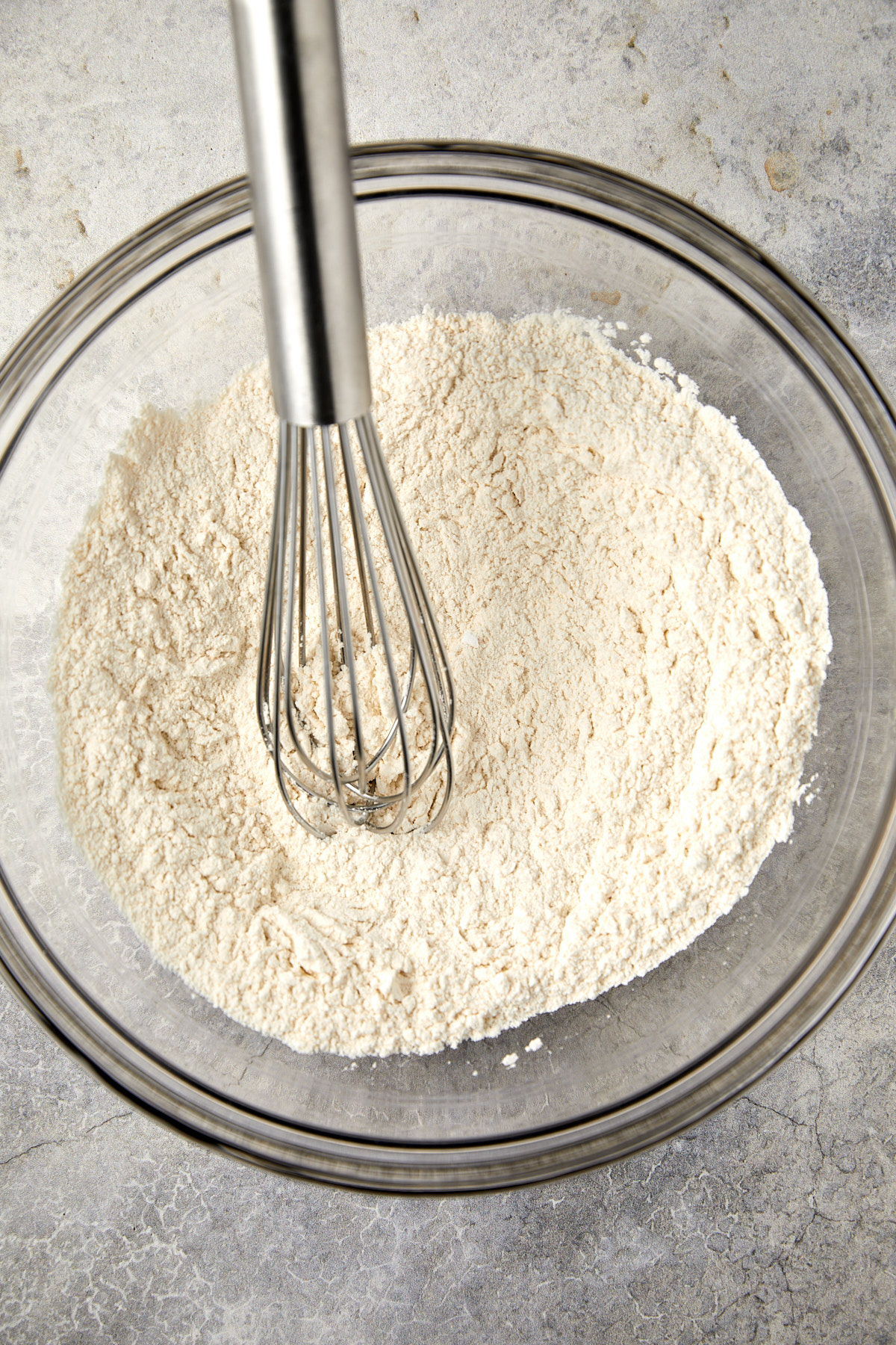 The image displays a clear glass mixing bowl on a gray surface, containing flour, baking powder, baking soda, and salt with a whisk. 