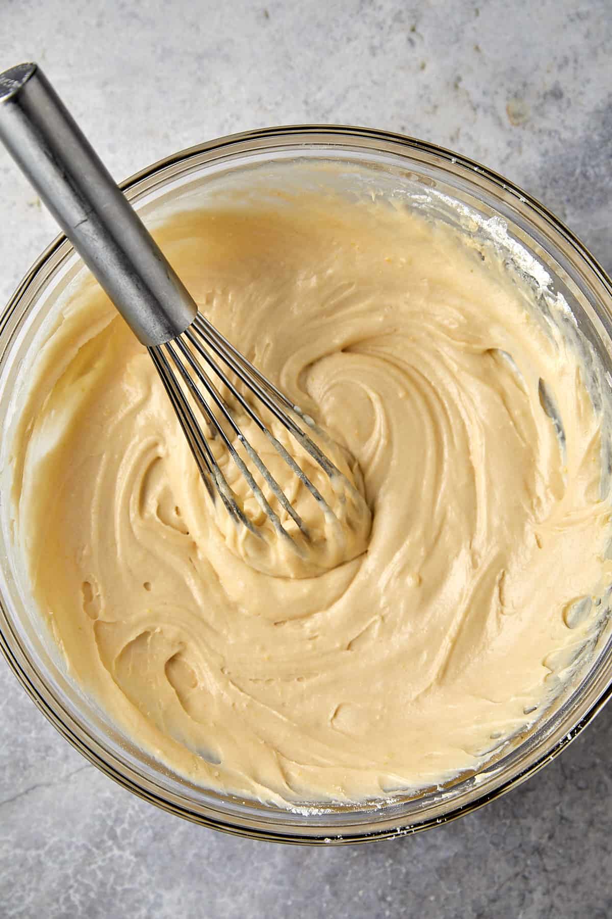 The image displays a glass mixing bowl containing a smooth, creamy batter. A metal whisk is partially immersed in the batter, indicating that the mixing process is either ongoing or just completed. The batter appears well-blended and is likely ready for the next step in a baking recipe, such as being poured into a baking pan to make a cake or loaf. The gray surface beneath the bowl suggests a kitchen countertop.