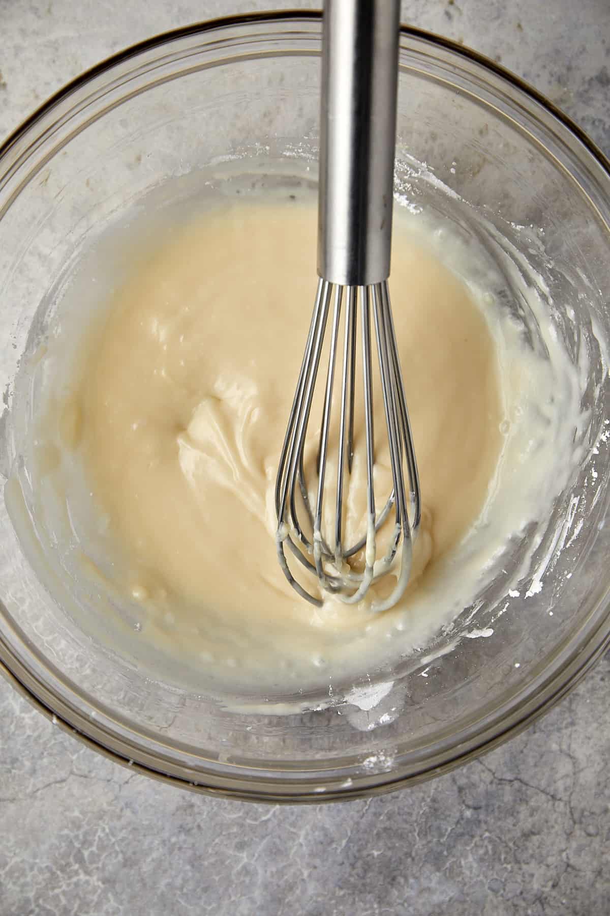 The image features a glass mixing bowl containing a pale, creamy white chocolate icing ready to put over the loaf cake. A metal whisk is submerged in the mixture. The consistency appears smooth and ready for use. 