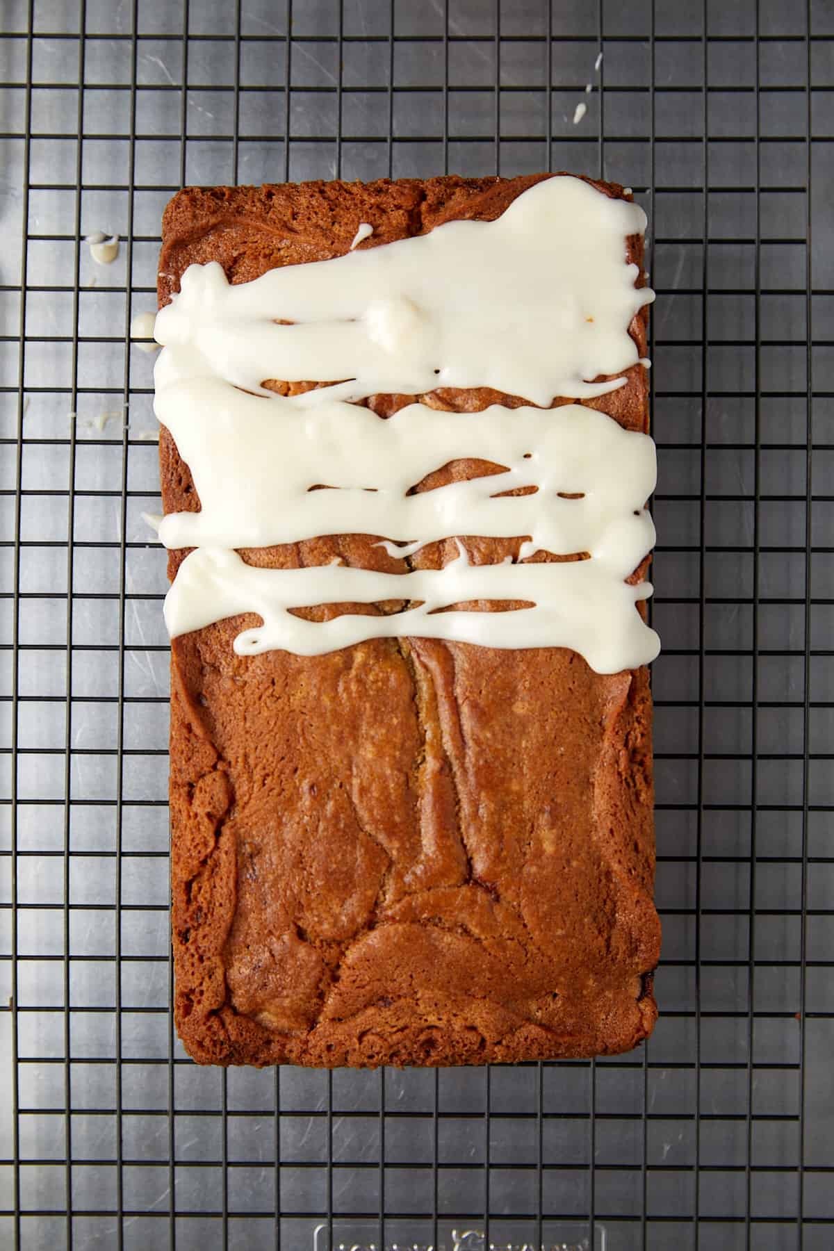 The image displays the raspberry white chocolate loaf cake with a rich, brown crust, presented on a wire cooling rack with a square grid pattern. The cake has a thick layer of white icing drizzled over the top, covering approximately half of its surface. The icing is uneven, with some areas thicker than others, creating an artistic, homemade appearance. The backdrop is a neutral gray, which might be a countertop, highlighting the contrast between the cake's warm tones and the icing's creamy white color.





