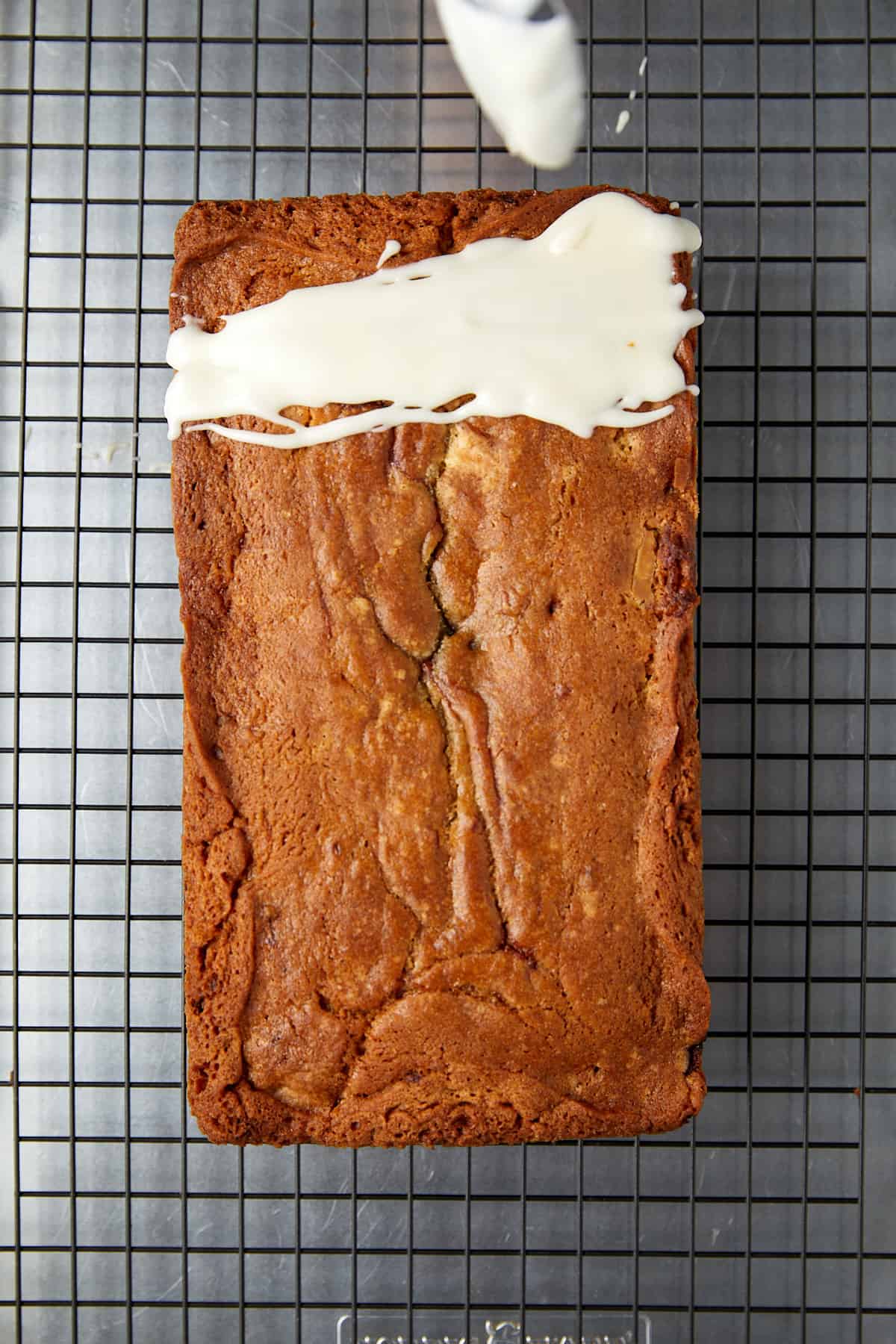 The image shows a golden-brown loaf cake with a cracked top, indicative of a well-baked treat. It rests on a wire cooling rack with a square grid pattern. A drizzle of white icing is being applied to the top of the cake, with the icing flowing down the sides in thick ribbons. The icing is likely made from a mixture similar to the one in the previous image, giving the cake a finished, tempting look. The background seems to be a gray surface, possibly a kitchen counter.