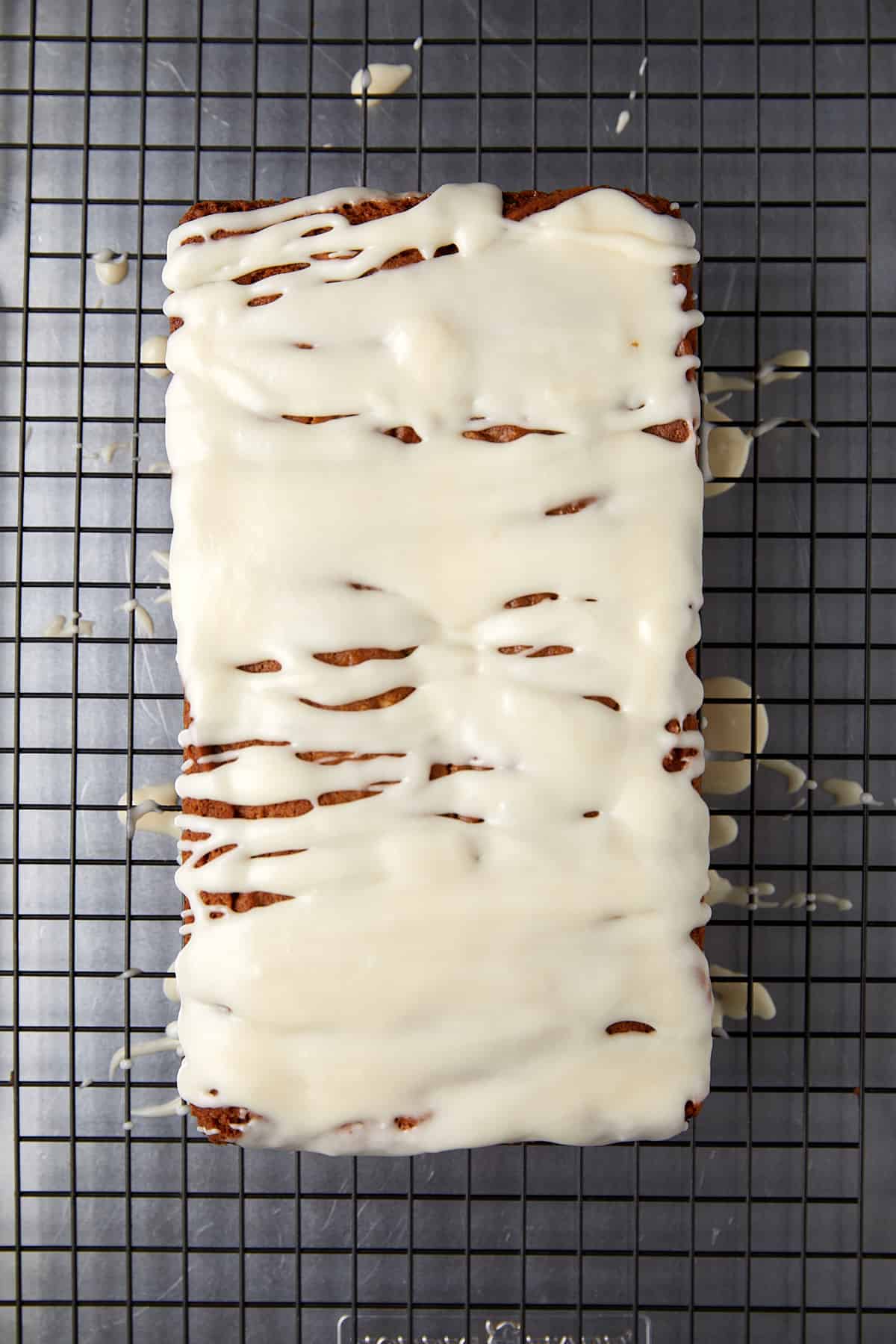 The image presents a loaf cake completely covered with a layer of white icing, placed on a wire cooling rack. The icing is spread across the top and has begun to drip down the sides, creating an appealing striped pattern that reveals parts of the cake's crust. The cooling rack's grid contrasts the smooth icing, and a few drops of icing have fallen onto the rack. The white chocolate raspberry loaf cake is now fully dressed and ready to be served, showcasing a finished bake that looks both homemade and delicious.






