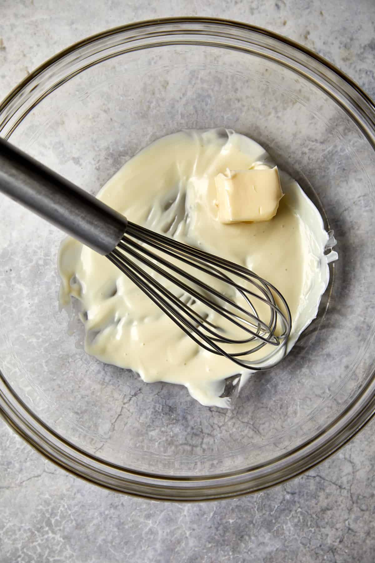The image displays a clear glass bowl with a mixture of melted white chocolate and a piece of solid butter. A metal whisk is also laying in the bowl.