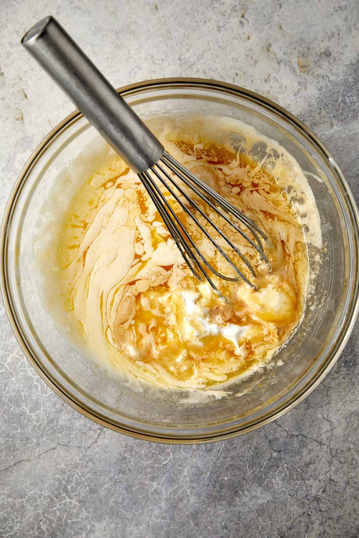 The image features a glass mixing bowl containing a mixture of the wet ingredients, vanilla, butter, and sour cream, in the process of being combined. A metal whisk is inserted into the mixture, highlighting the mixing action. 