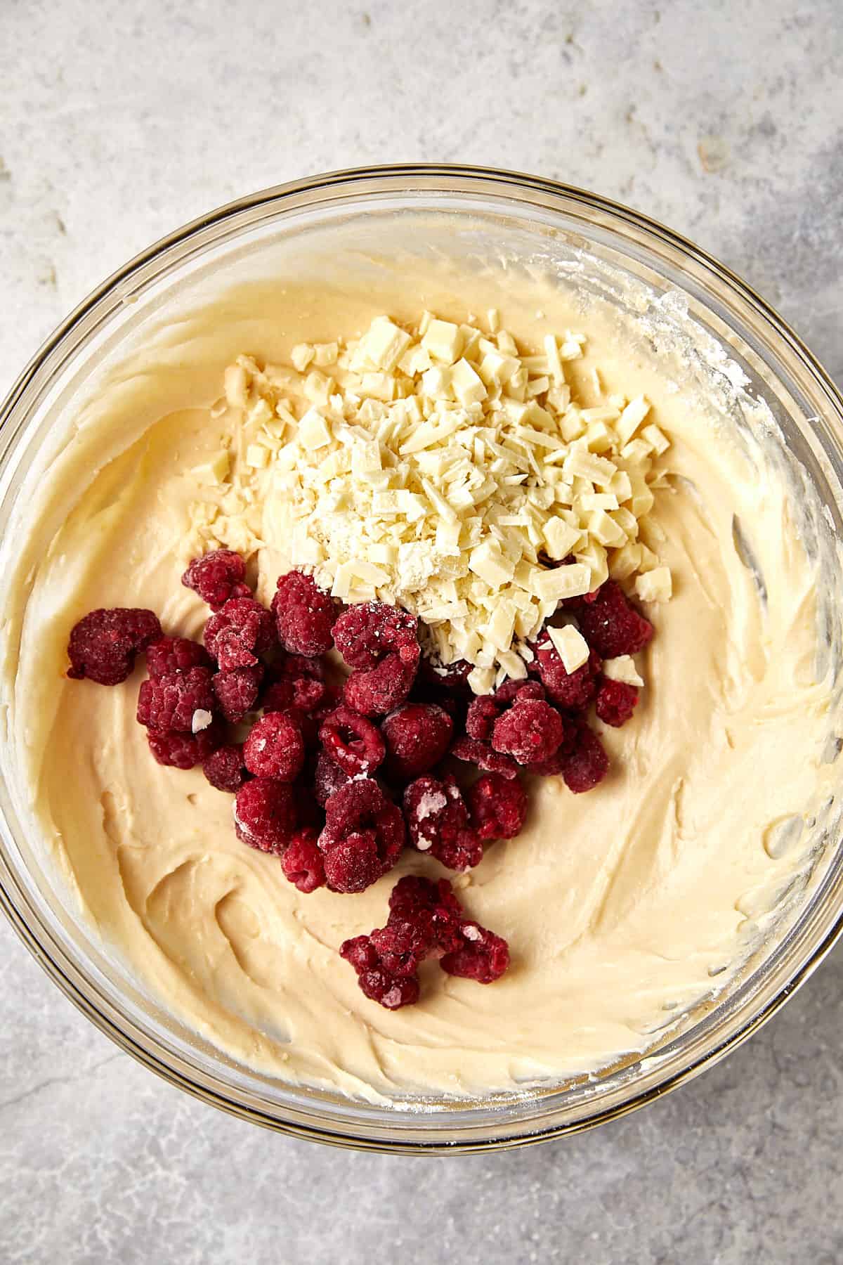 The image shows a glass mixing bowl filled with a smooth, creamy batter. On top of the batter, there is a generous amount of chopped white chocolate and a cluster of red raspberries. The ingredients have not yet been stirred into the batter. 