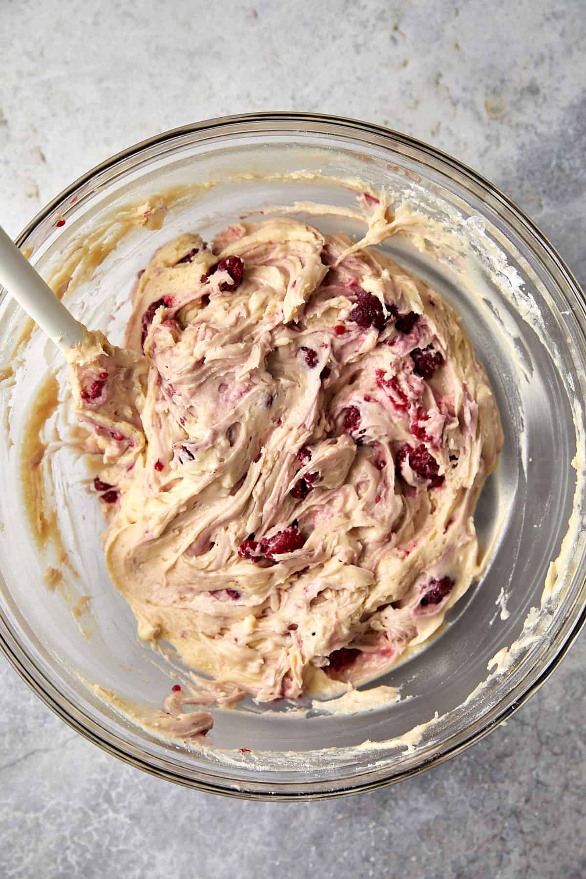 The image shows a glass mixing bowl with a batter that has raspberries and white chocolate folded into it. The berries and chocolate pieces are distributed throughout the creamy mixture, creating swirls of red and white. There is a spatula inside the bowl. 