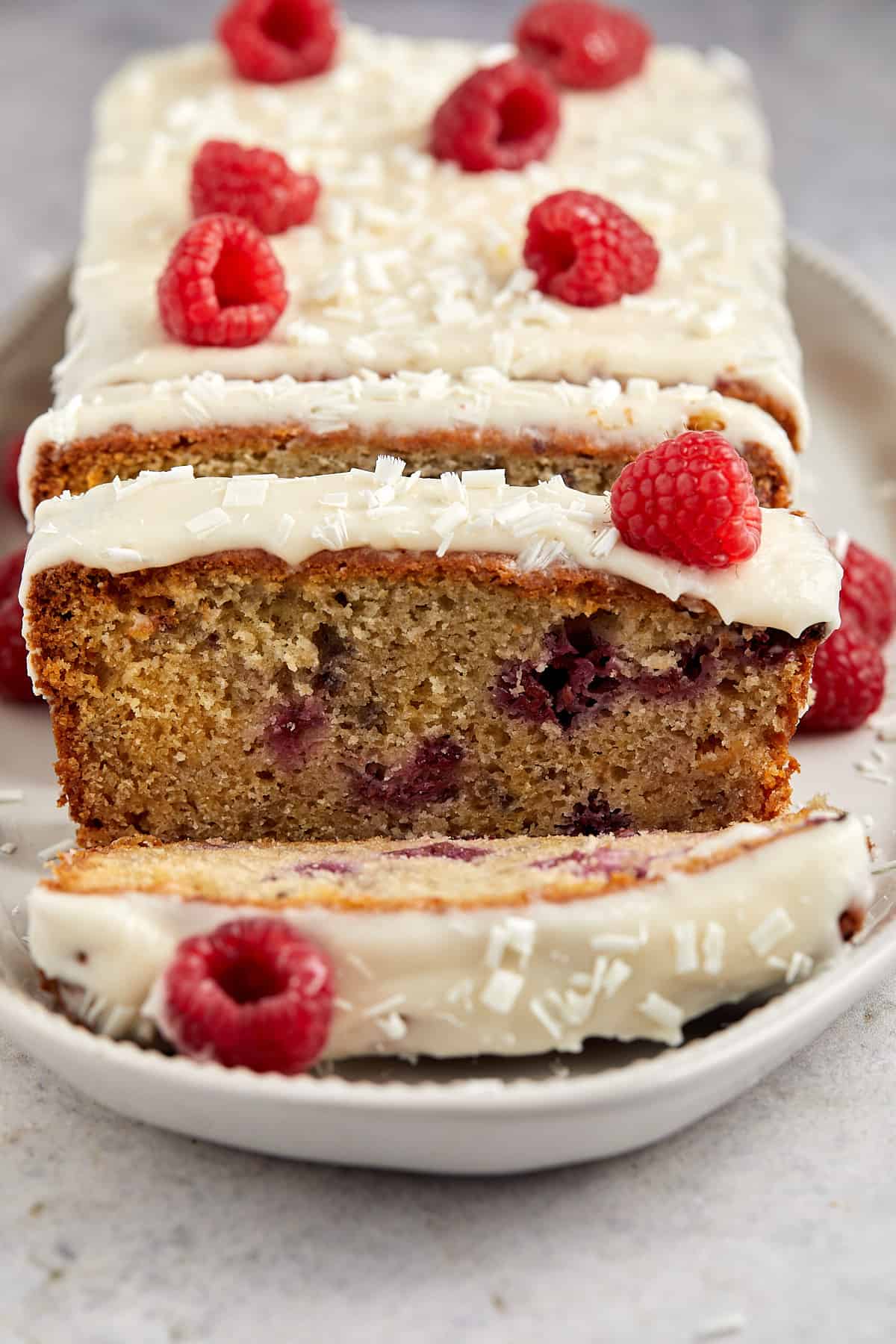 The image features a sliced raspberry and white chocolate loaf cake with white icing on top, garnished with fresh raspberries and sprinkled with white chocolate shavings. One slice is partially pulled out, revealing the moist interior of the cake studded with raspberries. The cake is placed on a light-colored plate, which sits on a textured surface. The photo captures the appealing texture and color contrast between the cake, the creamy icing, and the bright red raspberries.





