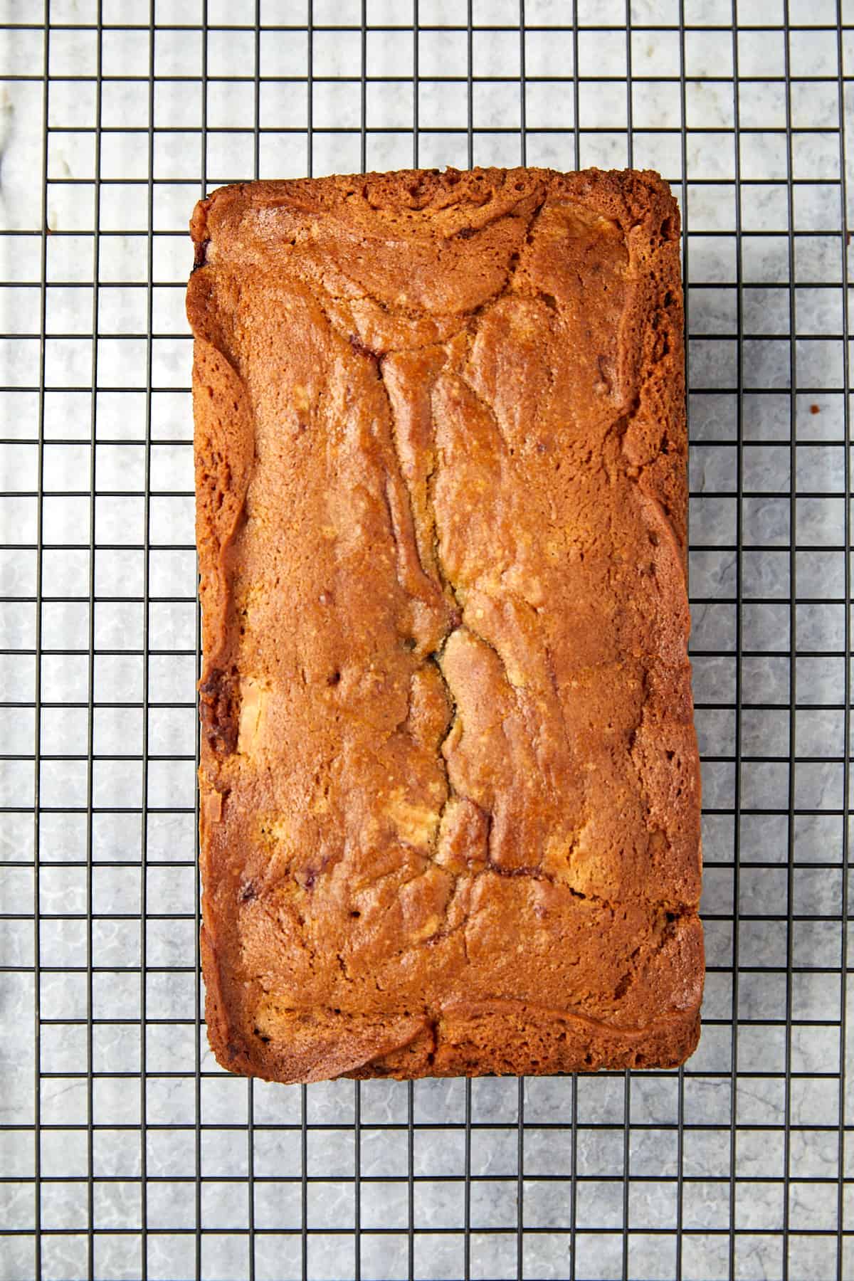 The image shows a freshly baked loaf cake with a golden-brown crust, cooling on a wire rack. The rack has a square pattern and is set against a light-colored background. The top of the loaf has a slight crack down the middle, characteristic of well-risen baked goods. 