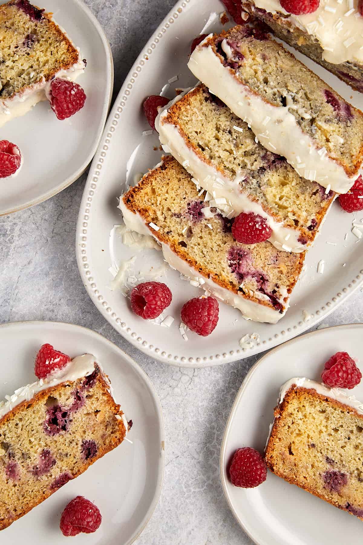 Overhead view of sliced raspberry and white chocolate loaf cake arranged on white plates, garnished with fresh raspberries and flakes of white chocolate. The plates are set on a grey marble countertop.