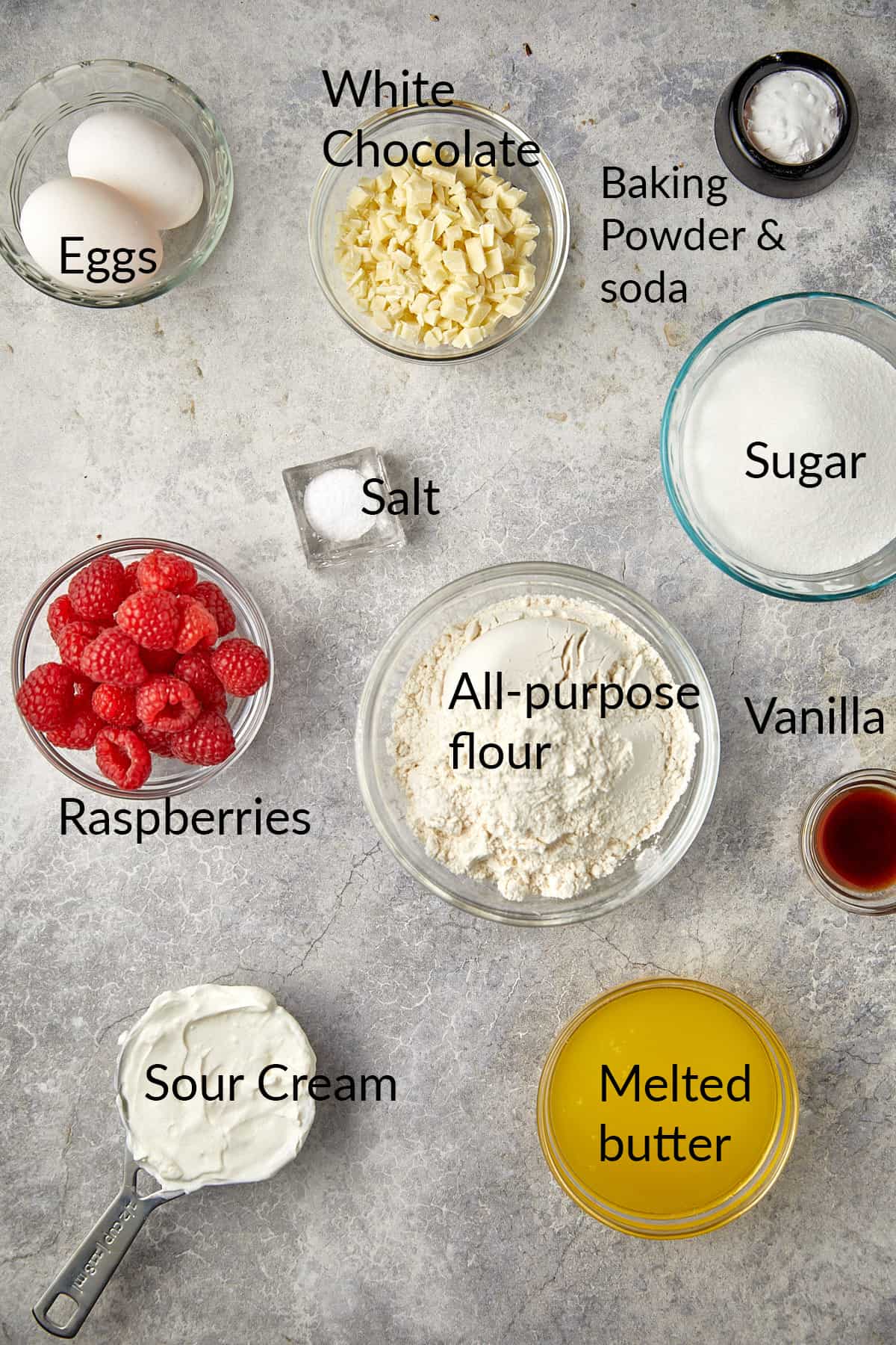 The image shows a collection of ingredients laid out on a gray surface, each labeled with its name. There are two eggs, a bowl of chopped white chocolate, a small container with baking powder and baking soda, a bowl of sugar, a pinch of salt, a bowl of fresh raspberries, a bowl of all-purpose flour, a small container of vanilla extract, a measuring cup with sour cream, and a bowl of melted butter. 