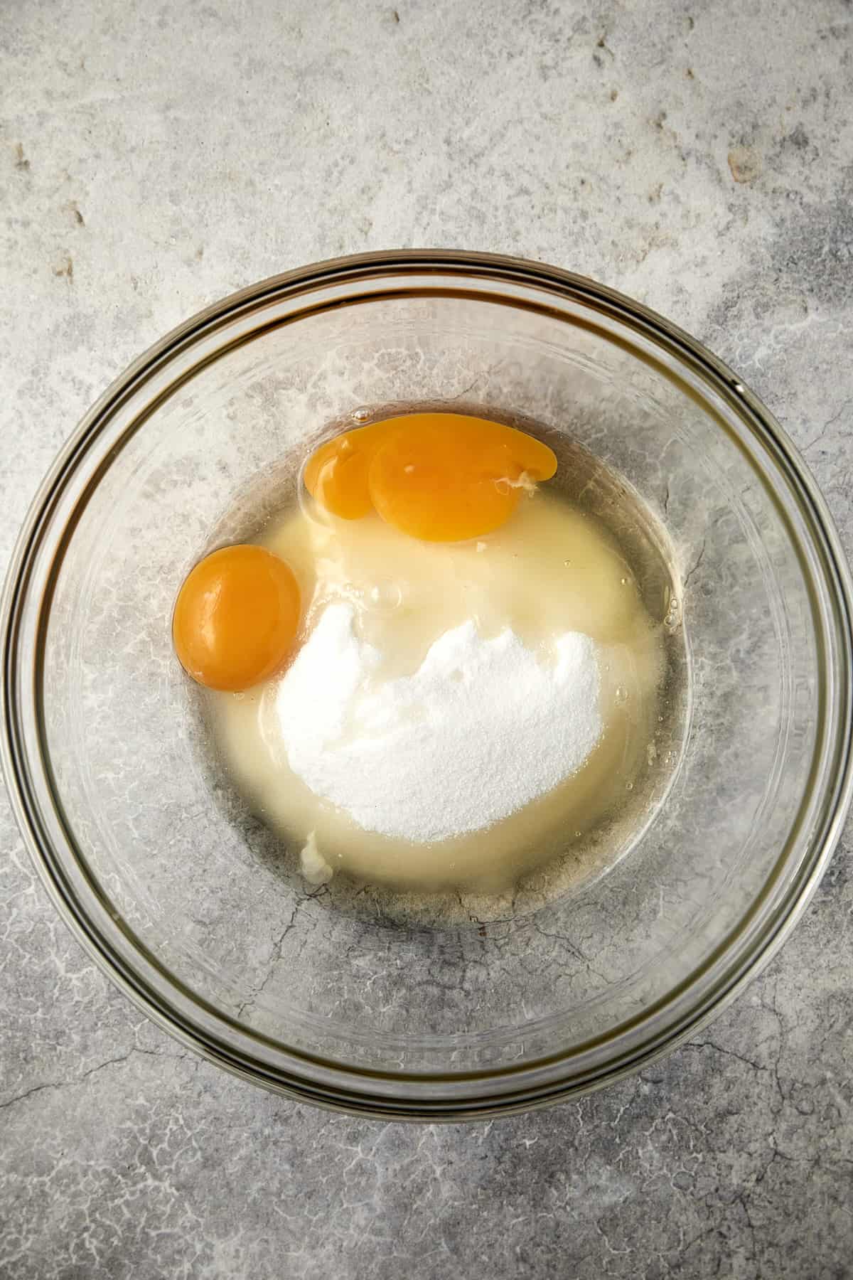 The image shows a clear glass bowl on a gray surface with two whole eggs and a pile of granulated sugar inside it. The ingredients are unblended.
