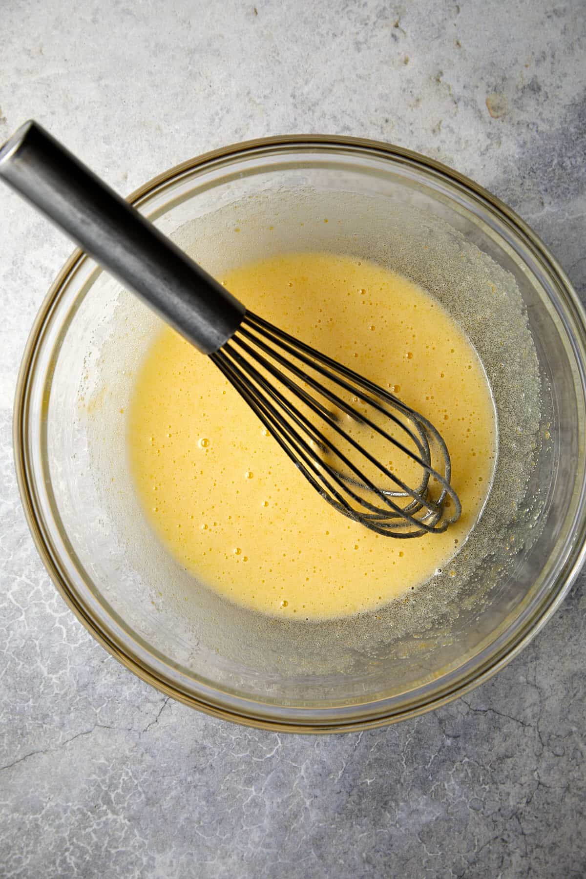The image depicts a clear glass mixing bowl on a gray surface containing a well-mixed combination of eggs and sugar. A metal whisk is in the bowl. 