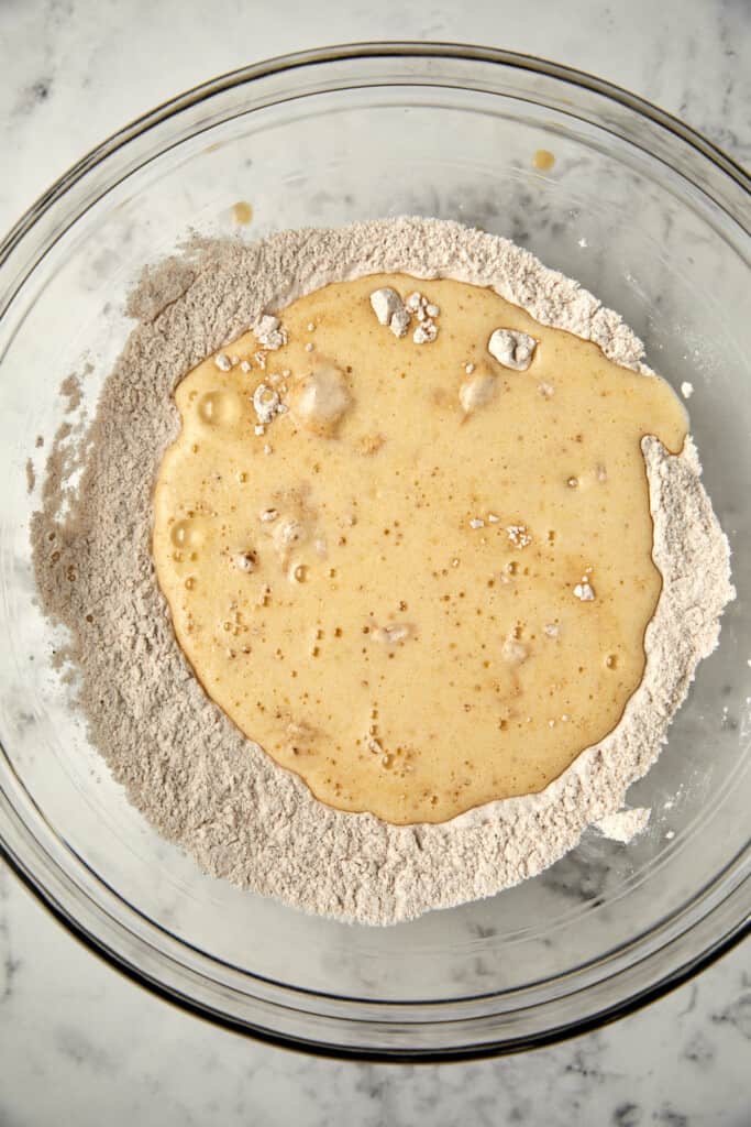 The image features a clear glass bowl on a marble surface containing dry ingredients, flour and leavening agents, with a wet batter mixture being poured over it. The batter appears to have a smooth texture with bubbles on the surface, suggesting it may have been recently whisked. The angle of the shot is from above, providing a detailed view of the ingredients and the initial stages of combining them, a common step in baking.