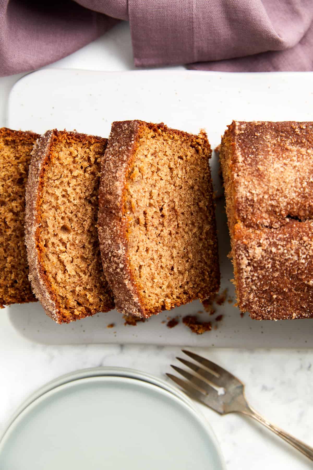 Looking down at the apple cider loaf cake sliced on a white board, plates and a fork are placed in front of loaf.