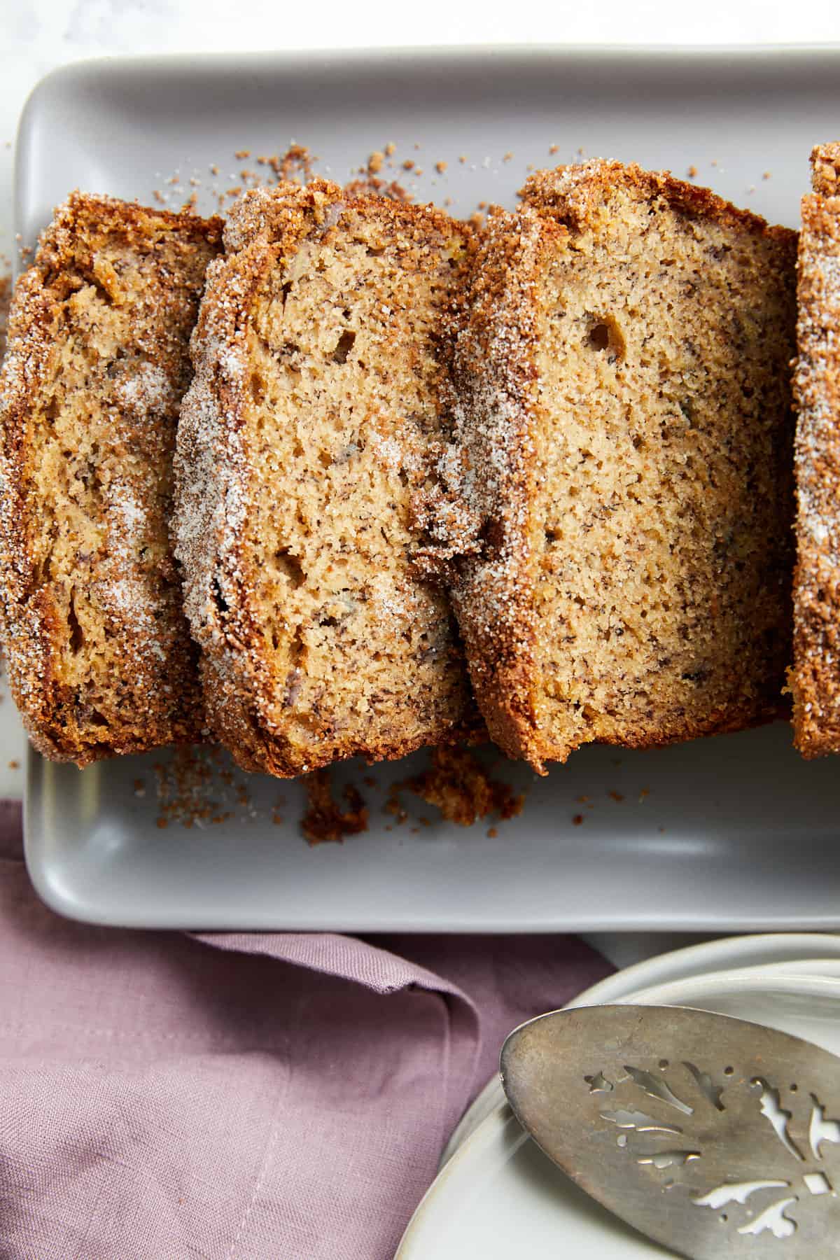 Looking down and cinnamon crunch bread sliced on a gray platter with plates and a server utensil sitting next to it.