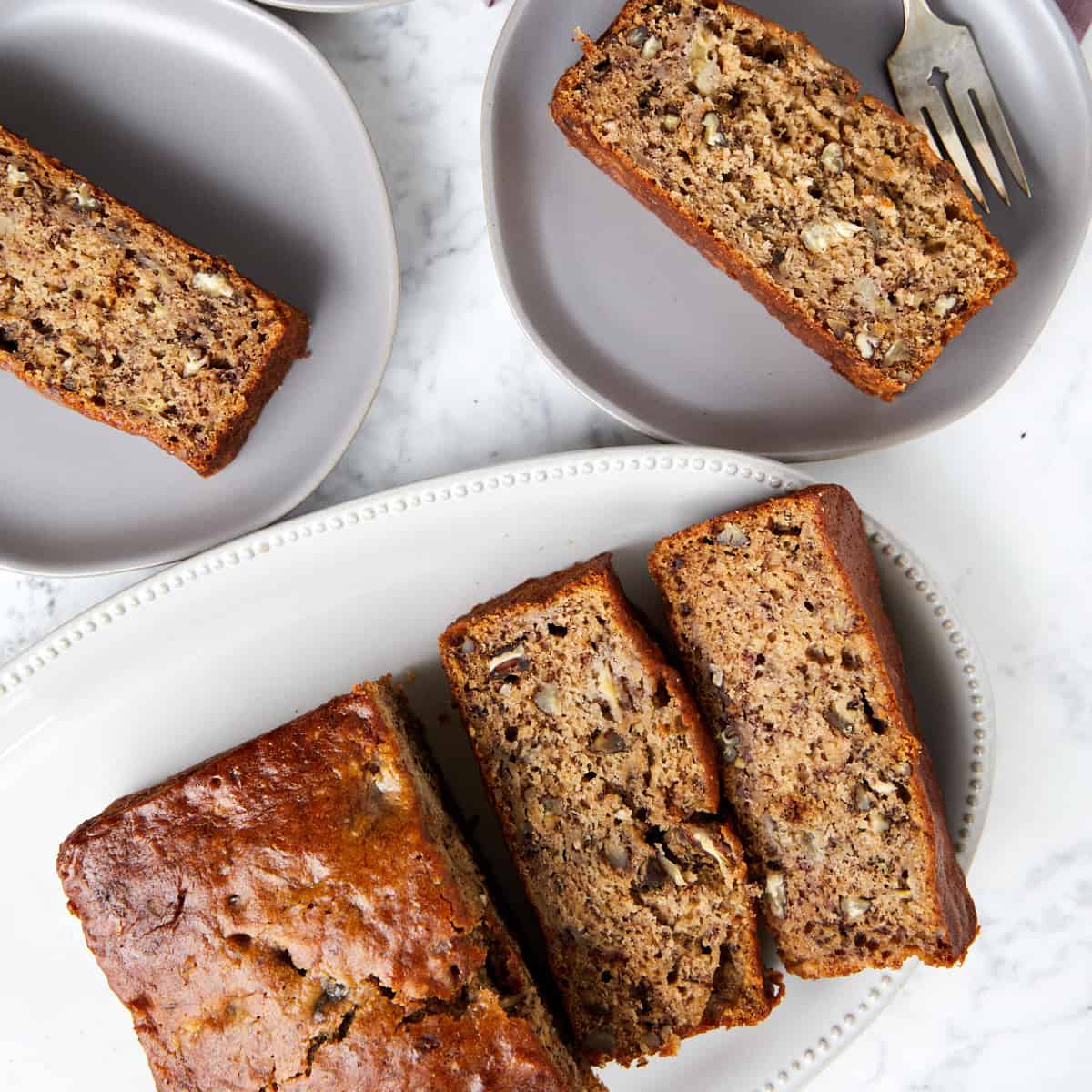 looking down at baked banana bread with pecans sliced on plates with a fork next to a piece of bread on one plate.