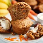 Baked banana carrot muffins sitting on a white plate. A slice of one-half of a muffin is sitting on top of a whole muffin. Another muffin is split with butter on it laying on the sides of the whole muffin.