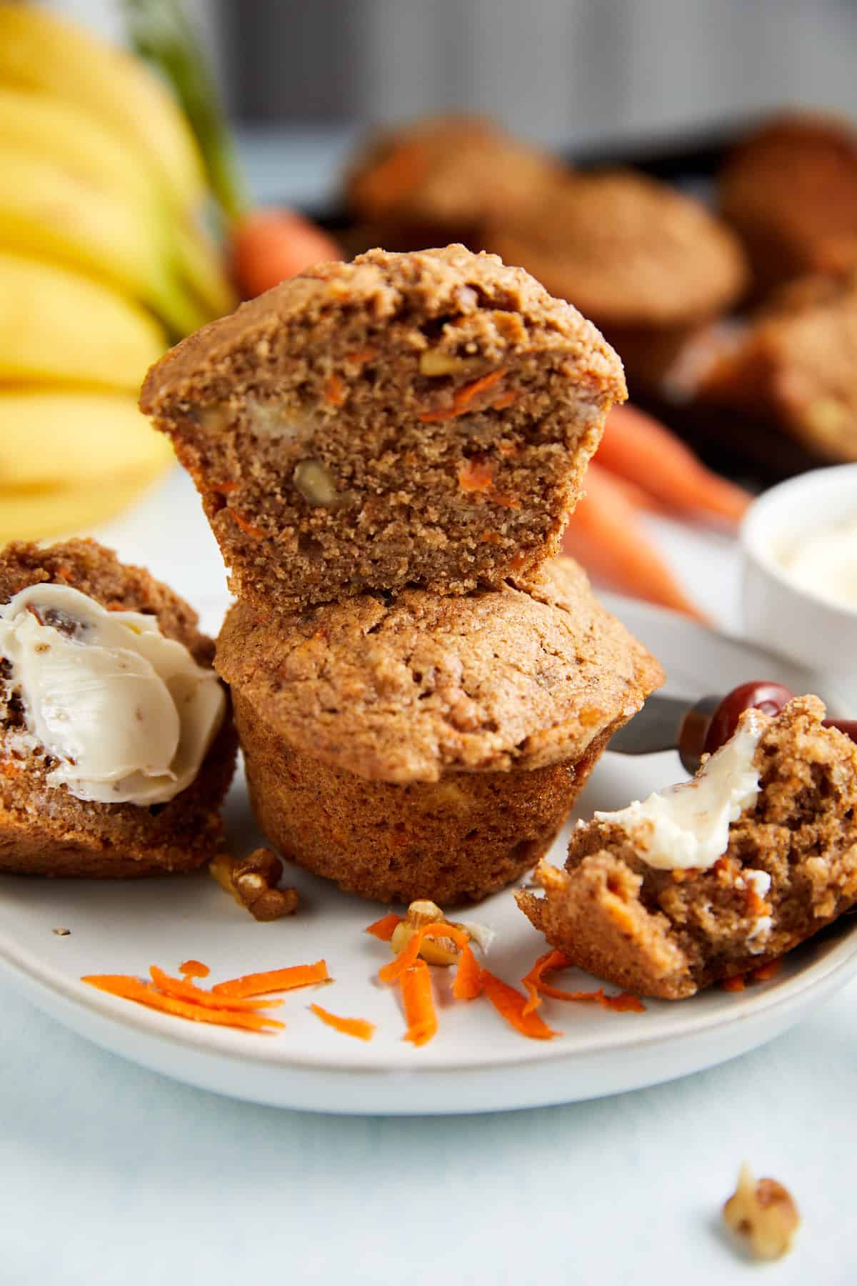 Baked banana carrot muffins sitting on a white plate. A slice of one-half of a muffin is sitting on top of a whole muffin. Another muffin is split with butter on it laying on the sides of the whole muffin.