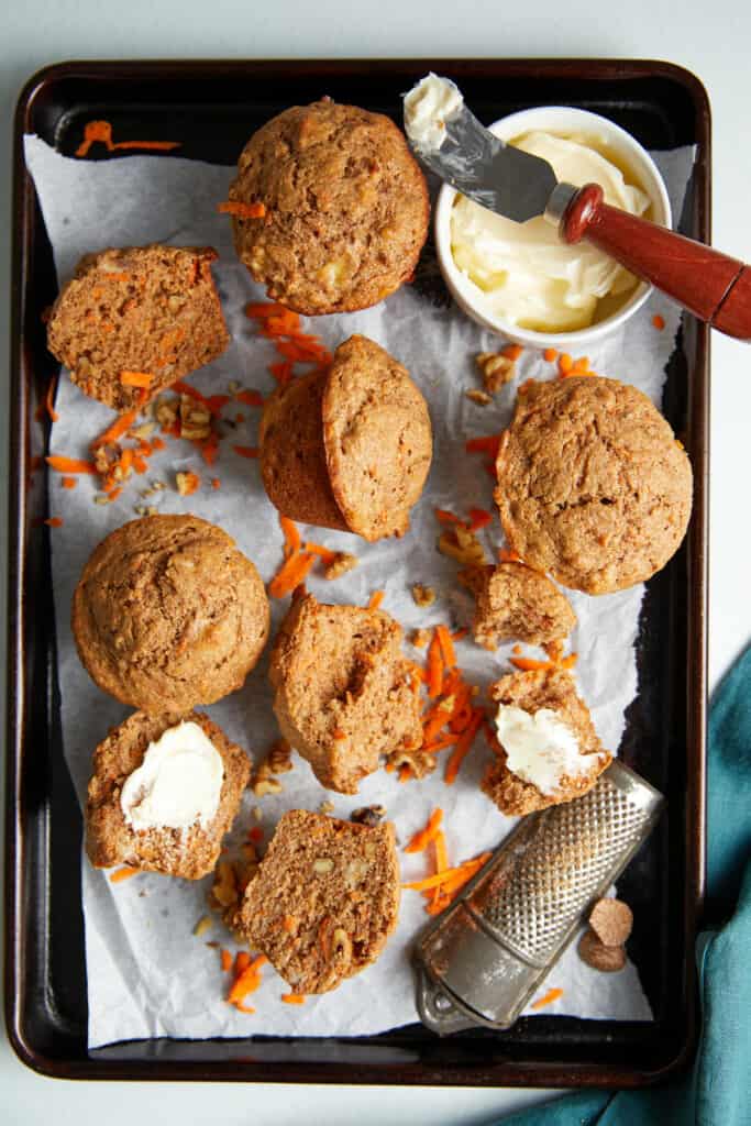 Baked banana carrot muffins on a cookie tray lined with white paper. Four whole and others are sliced in half. A white bowl of butter with a knife with a smear of butter on the knife sits on the bowl. In the lower left of the tray sits a old-time nutmeg grinder.