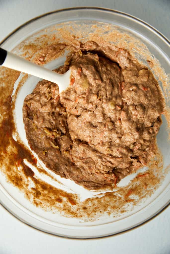 looking down at clear bowl with the carrots and walnuts being folded into wet ingredients with a spatula.