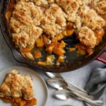 looking down at the cast iron pan of peach cobbler with two plates with servings of the cobbler and ice cream along three spoons.