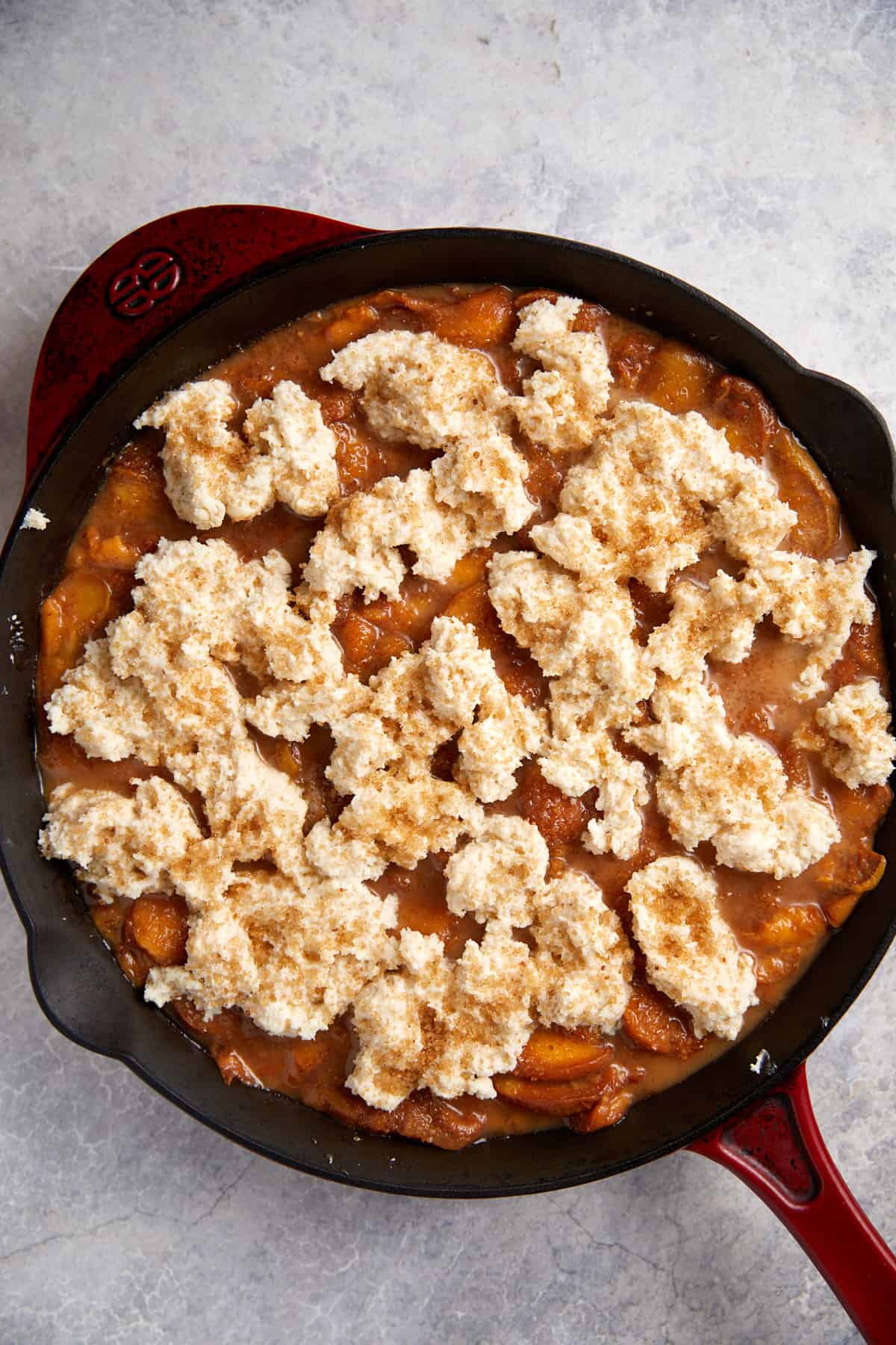looking down at cast iron peach cobbler with raw biscuit dough dotted over the peaches.
