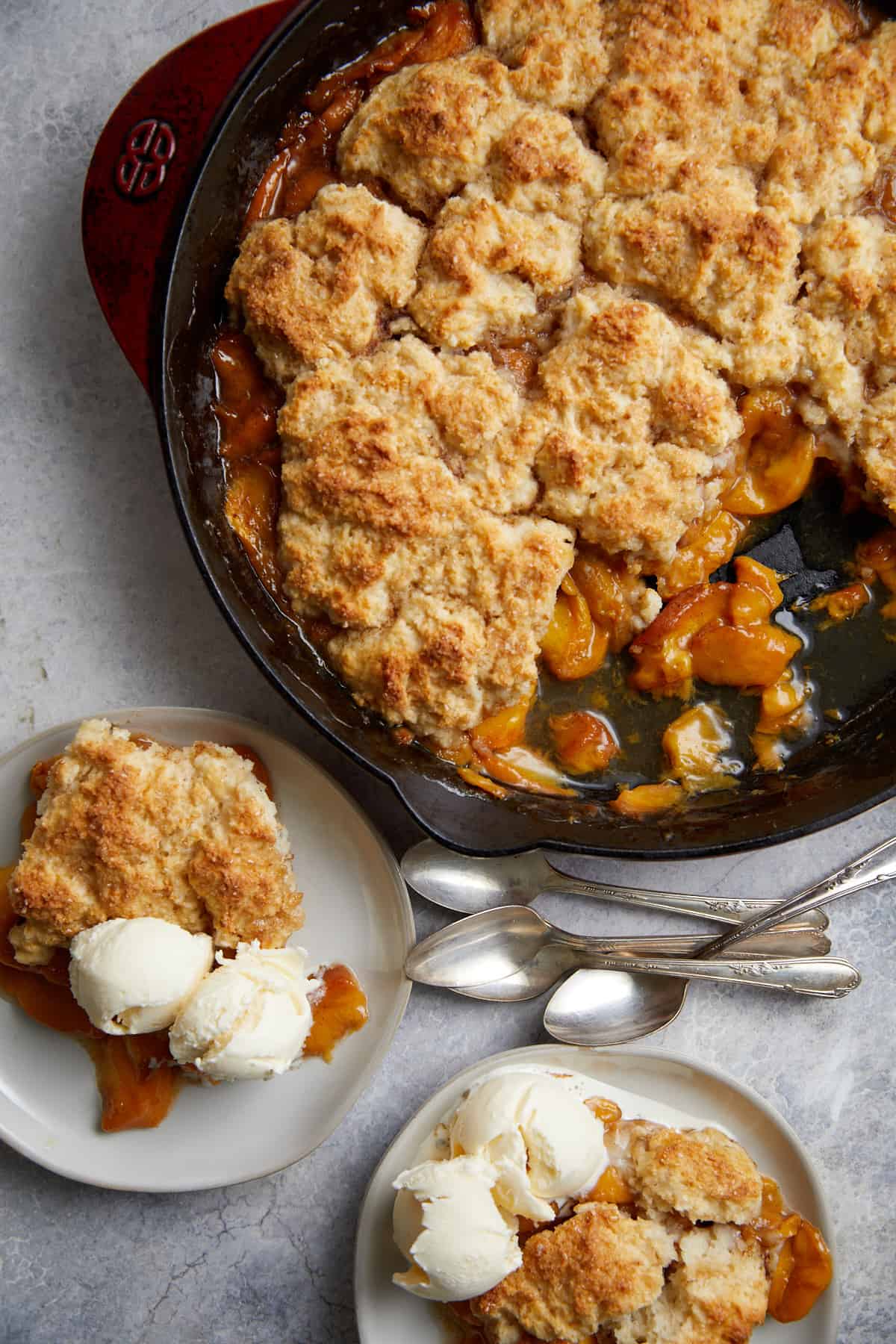 looking down at the cast iron pan of peach cobbler with two plates with servings of the cobbler and ice cream along with a pile of spoons.