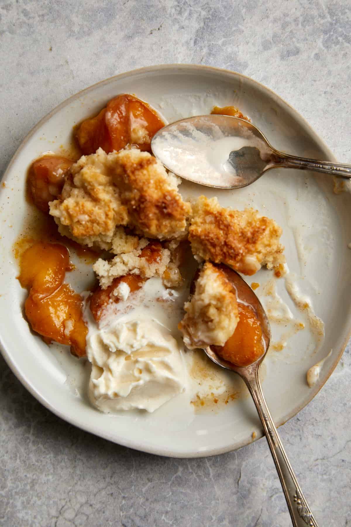 baked peach cobbler and vanilla ice cream on a white plate with two spoons.