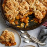 looking down at the cast iron pan of peach cobbler with two plates with servings of the cobbler and ice cream along with a pile of three spoons.
