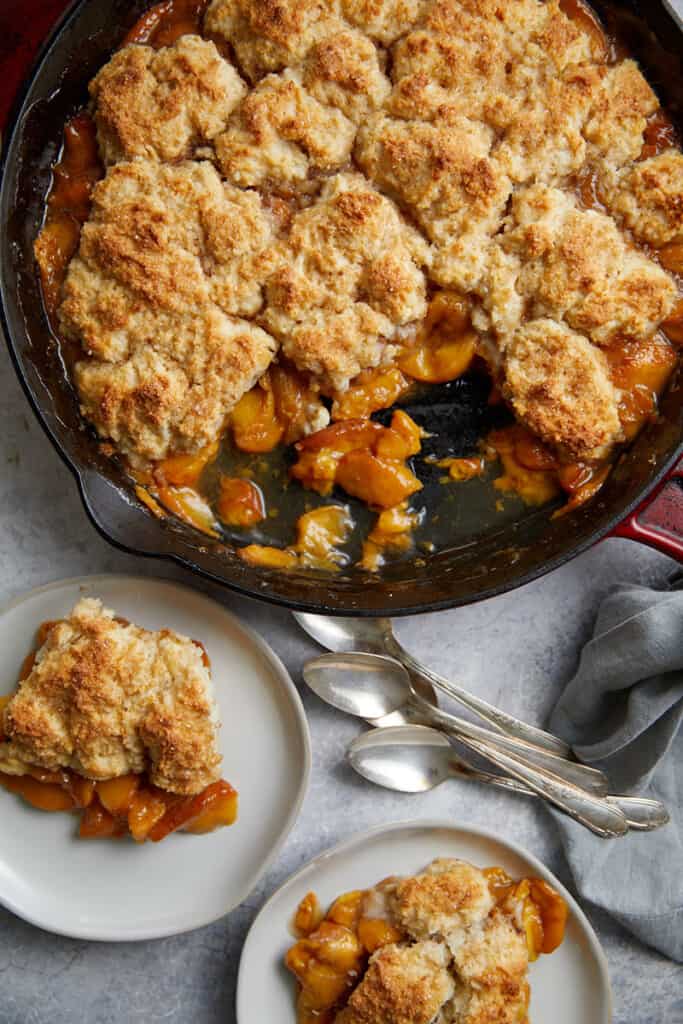 looking down at the cast iron pan of peach cobbler with two plates with servings of the cobbler and ice cream along with a pile of three spoons.