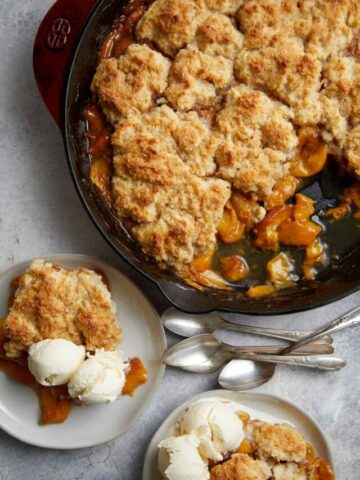 looking down at the cast iron pan of peach cobbler with two plates with servings of the cobbler and ice cream along with a pile of spoons.