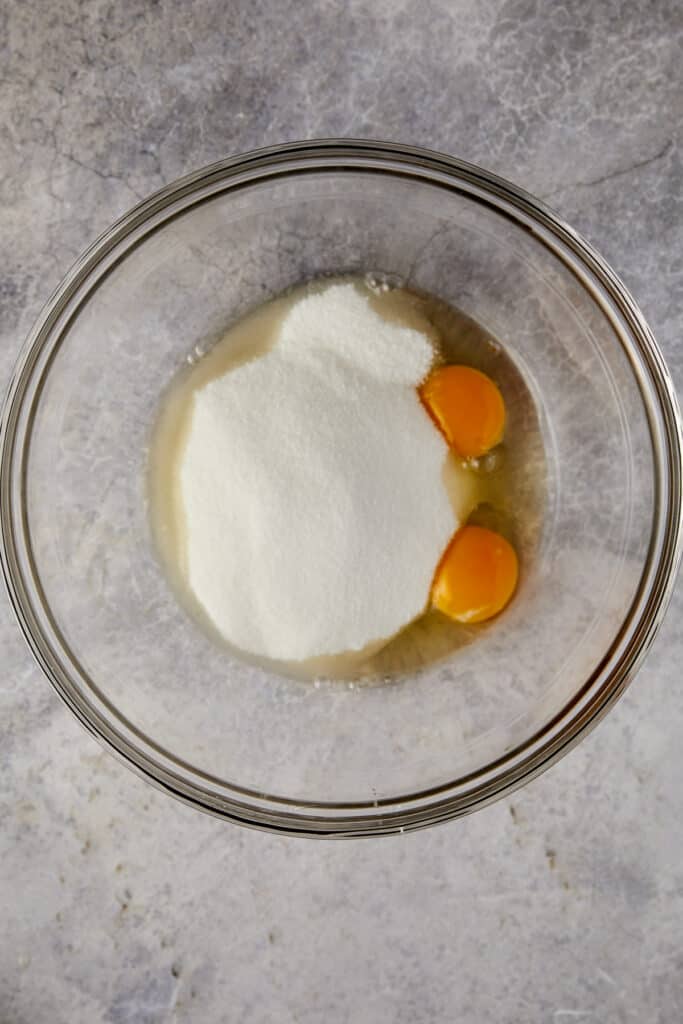 An overhead view of granulated sugar and eggs in a clear bowl.