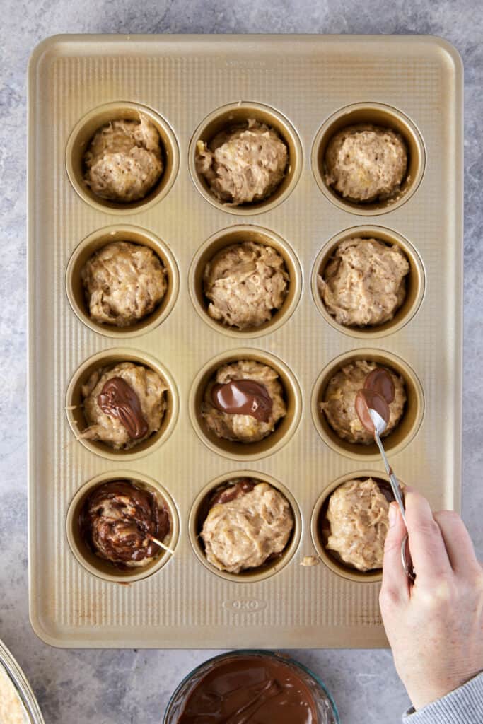 The overhead view of batter filled muffin tin with a teaspoon of pouring Nutella into the batter.