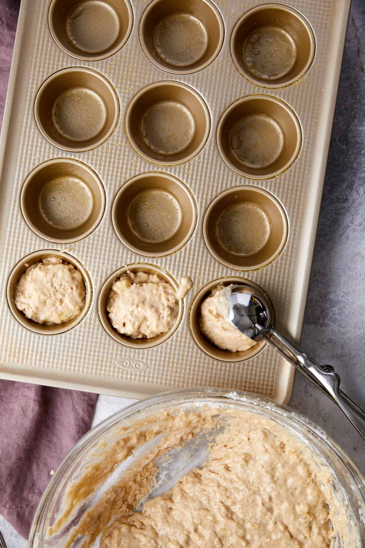 Overhead view of a muffin tin getting filled with cinnamon muffin batter using an ice cream scoop. 