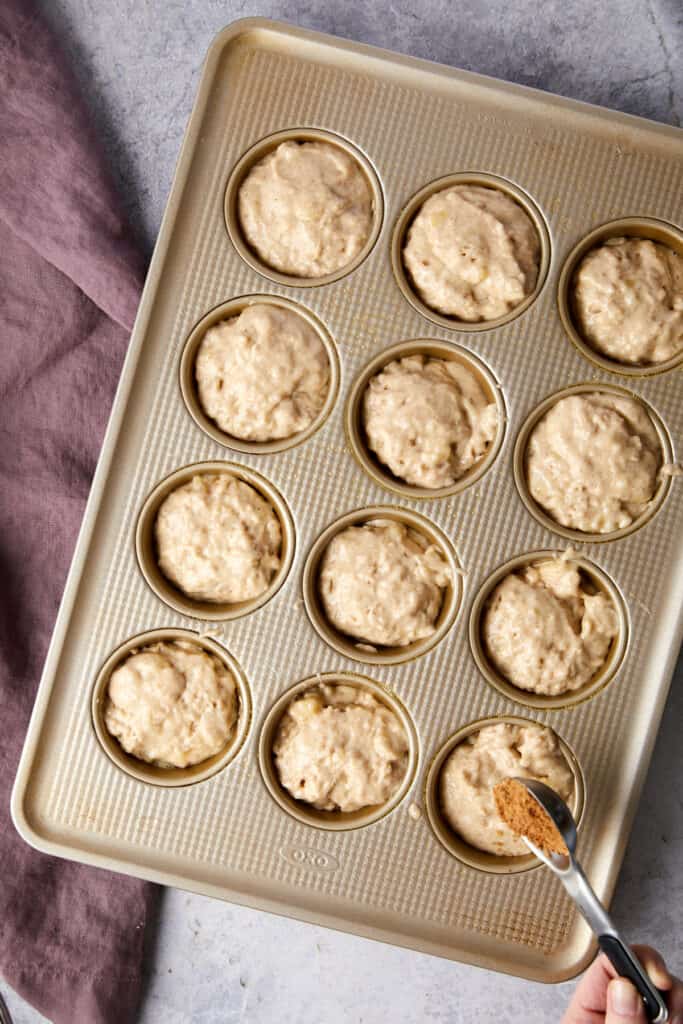 Overhead view of cinnamon swirl banana muffins batter in the muffin tin. A hand is pouring out a teaspoon of cinnamon sugar mixture on top of the batter.