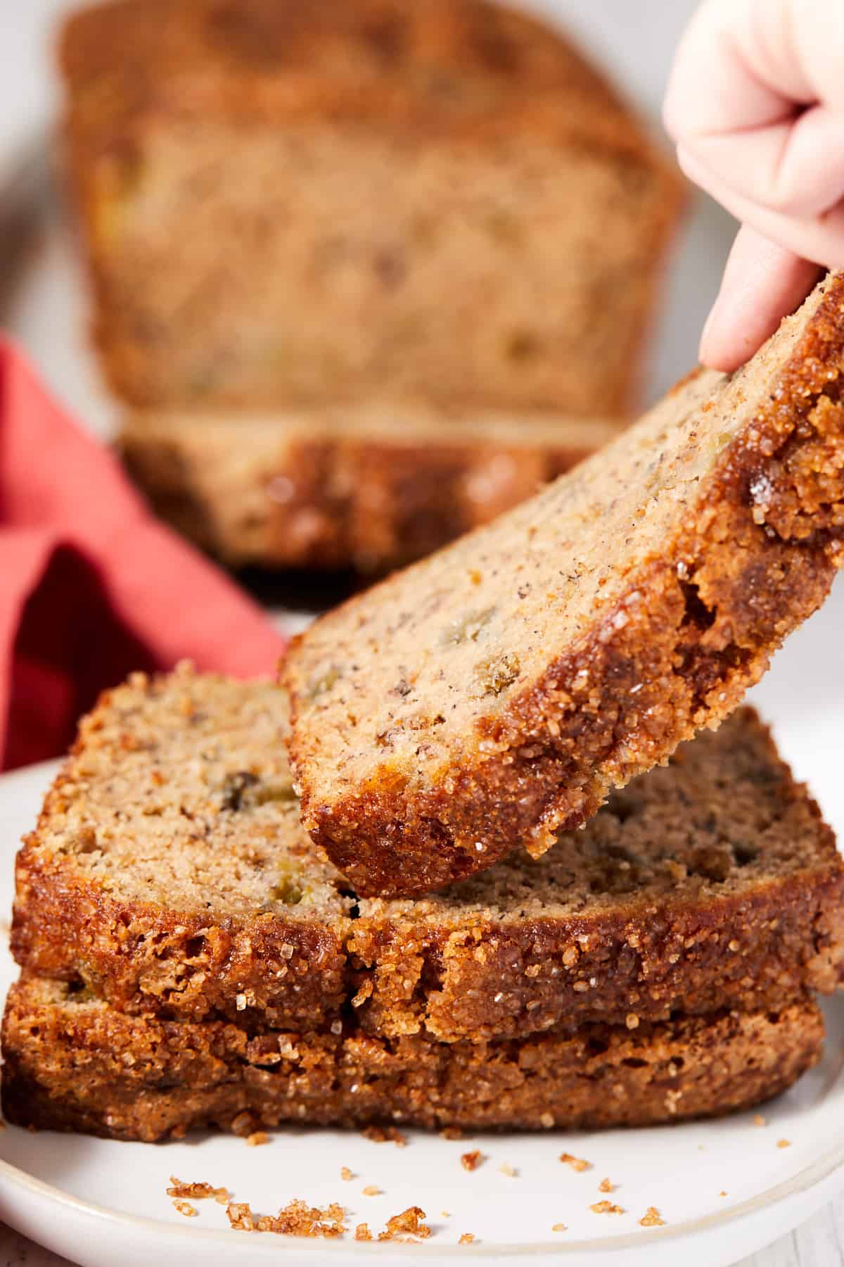 Three slices of Rhubarb banana bread are sitting on a plate with one slice being lifted by a hand.